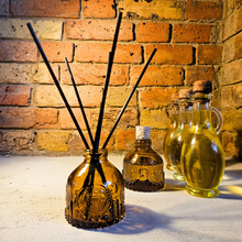 Load image into Gallery viewer, Luxury amber diffuser with black reeds in amber glass bottle on rustic surface against brick wall background