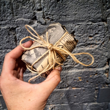 Load image into Gallery viewer, Hand holding English Sea Salt and Smoked Sage soy wax candle wrapped in vintage newspaper with twine bow against a textured dark wall background